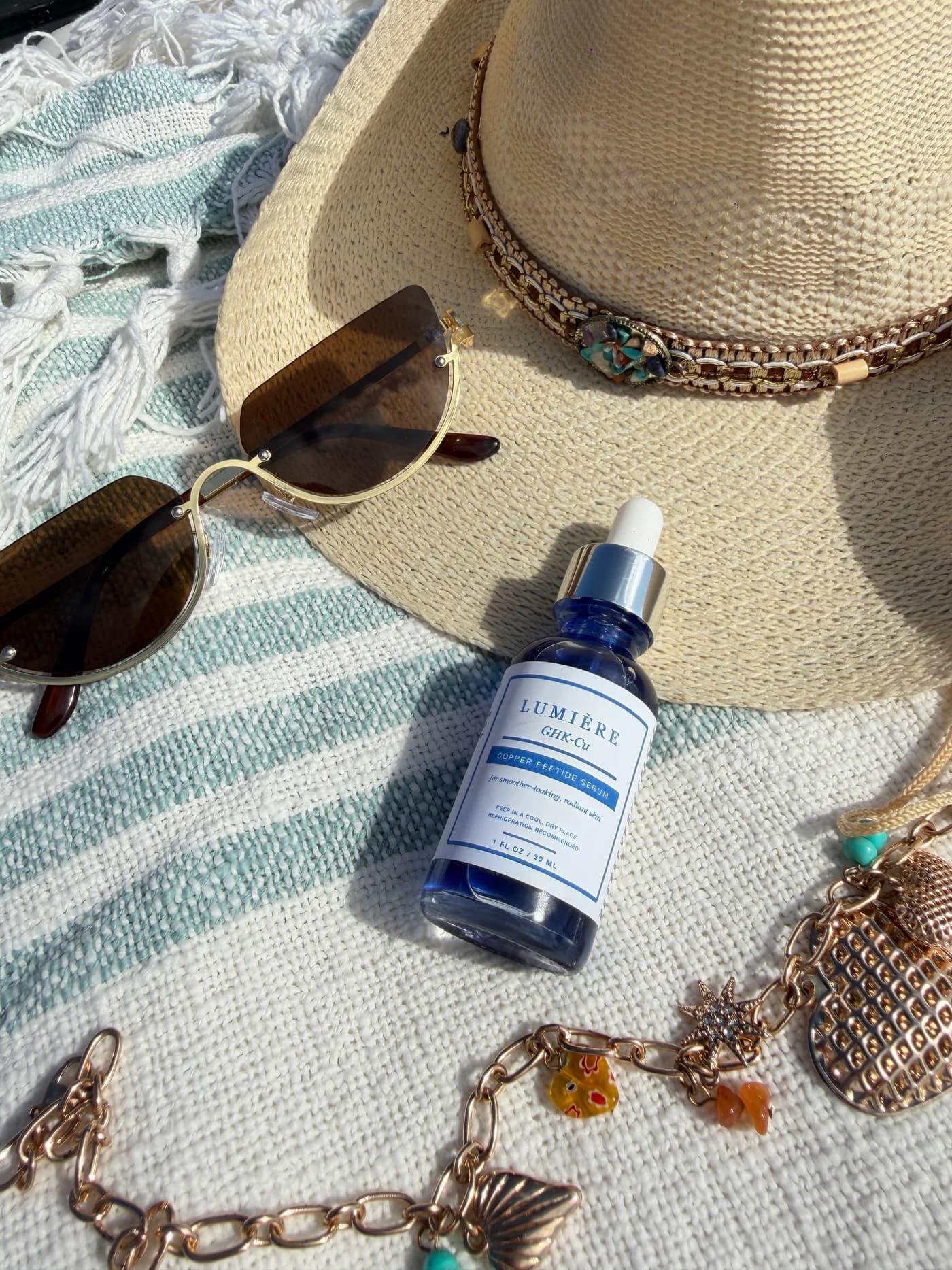 Lumiere serum on a striped beach towel beside a woven hat and sunglasses