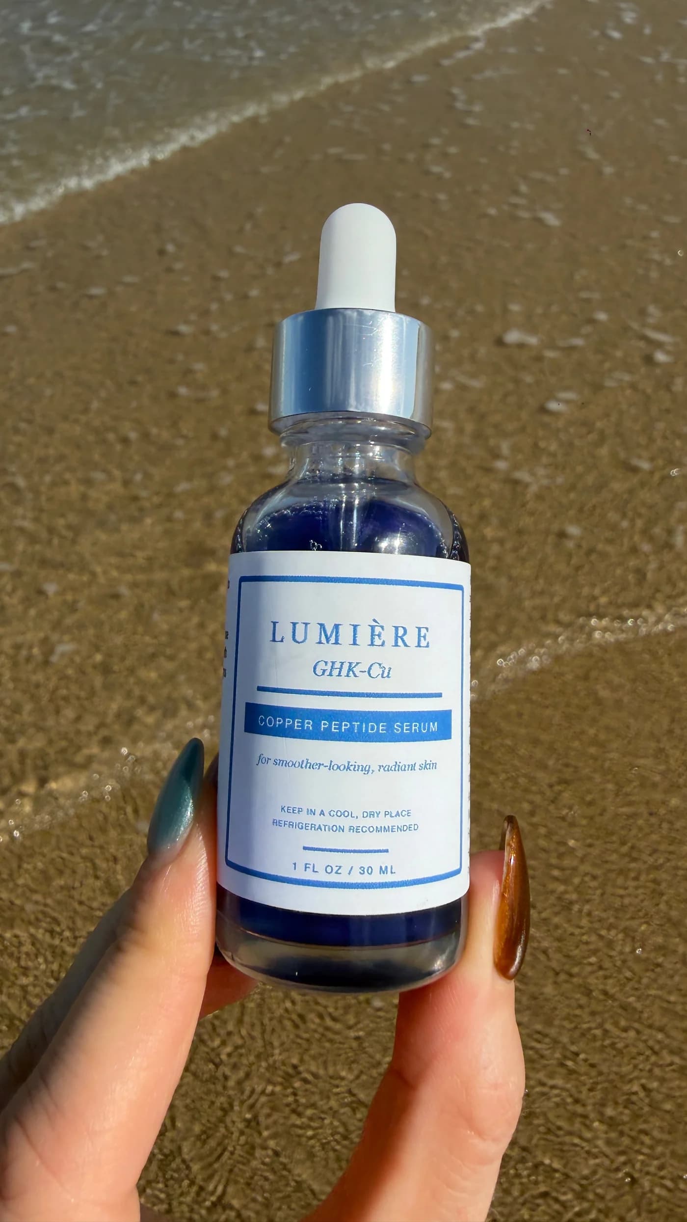 A hand holding the Lumiere serum bottle above the sand at the shoreline